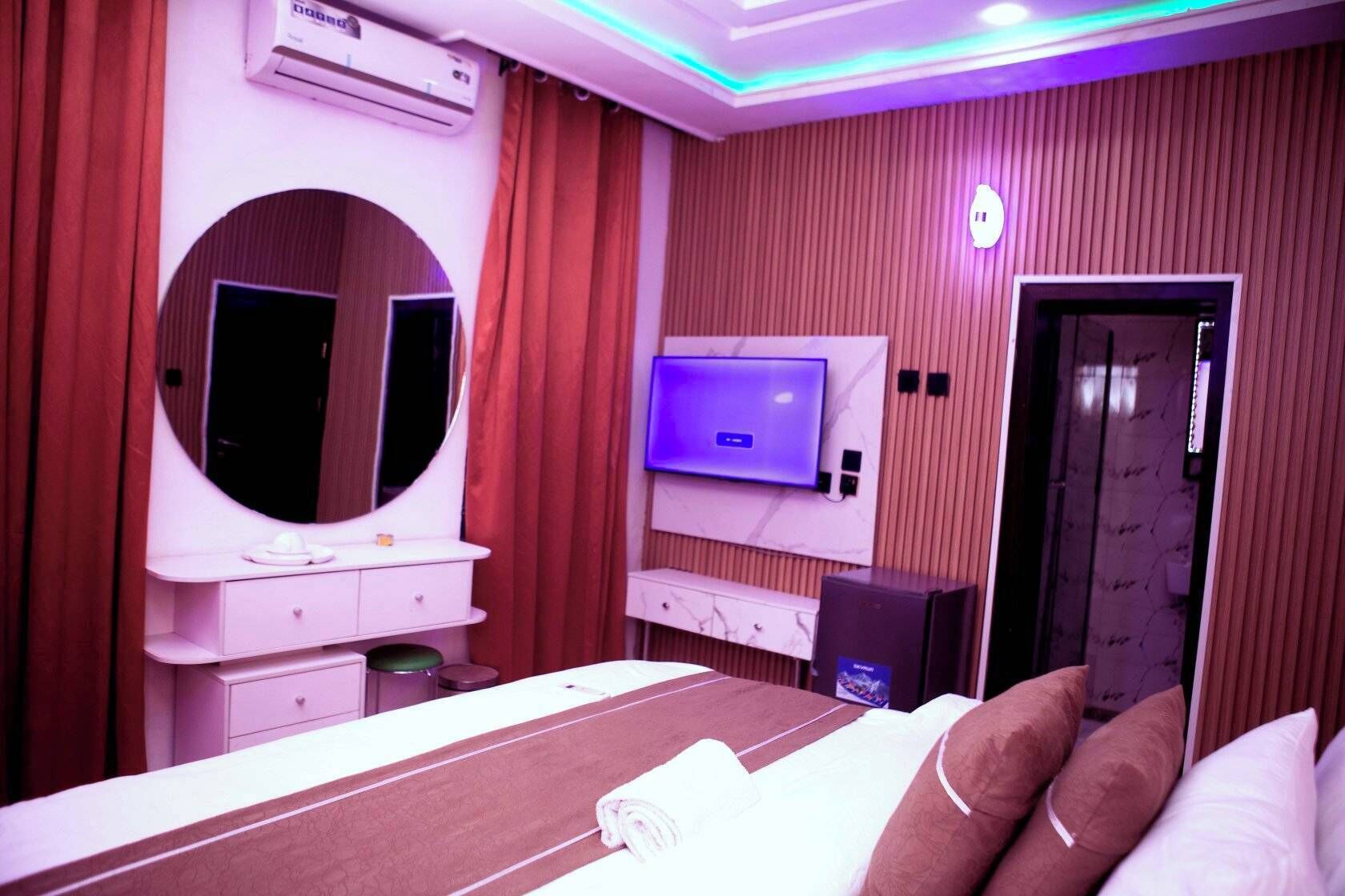 A hotel room with a bed, a vanity with a mirror, a television, and an air conditioning unit.