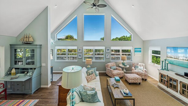 A spacious living room with a high ceiling, large windows, a ceiling fan, and a television.
