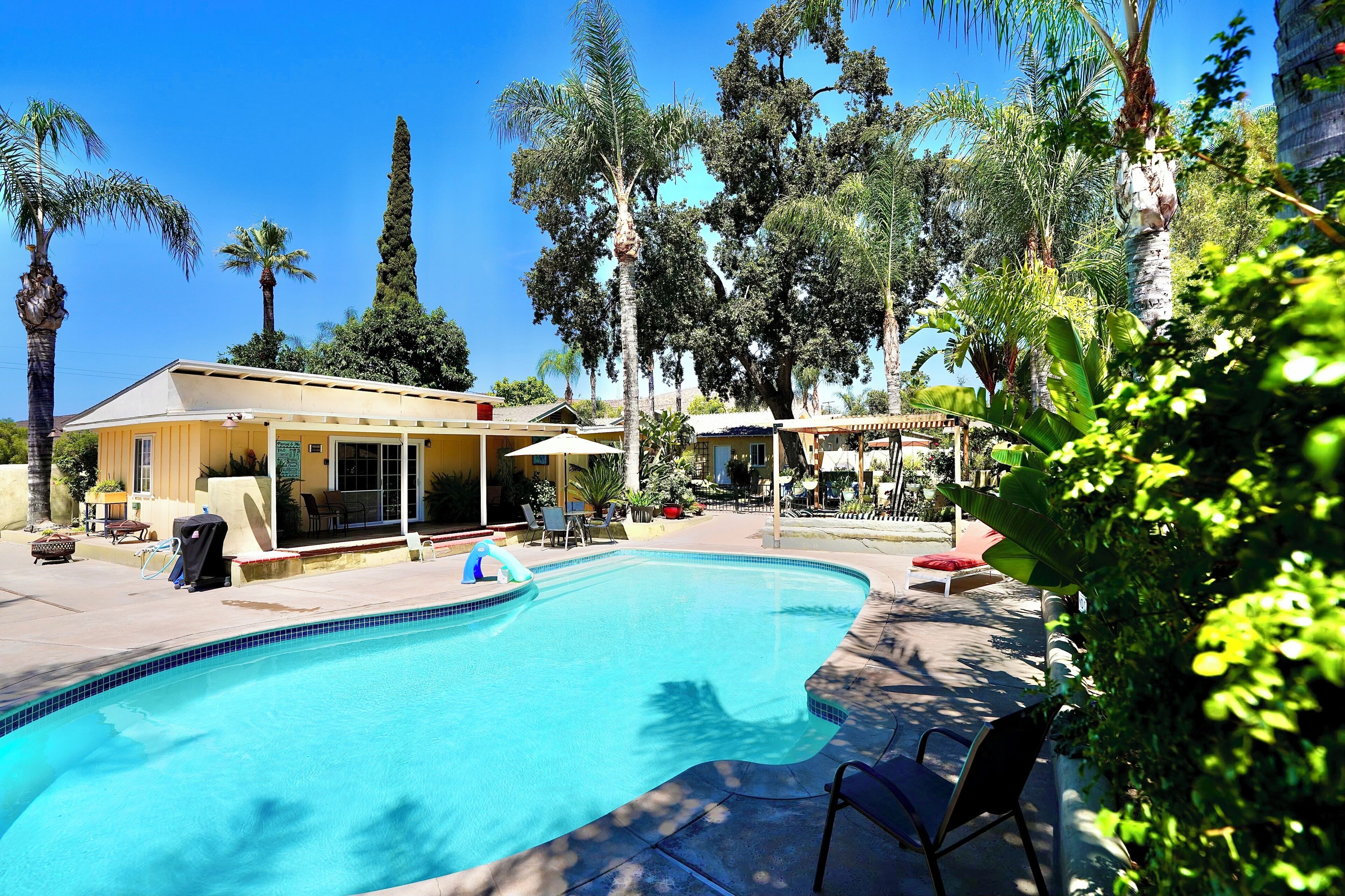 A swimming pool with lounge chairs and a covered seating area, surrounded by palm trees and a building with a yellow facade.