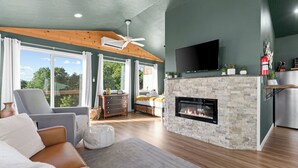 A modern living room with a fireplace, a flat-screen TV, and a large window overlooking greenery. - Views! Private Hot tub and Fire Pit! Lux Couple's Retreat (Omaha)