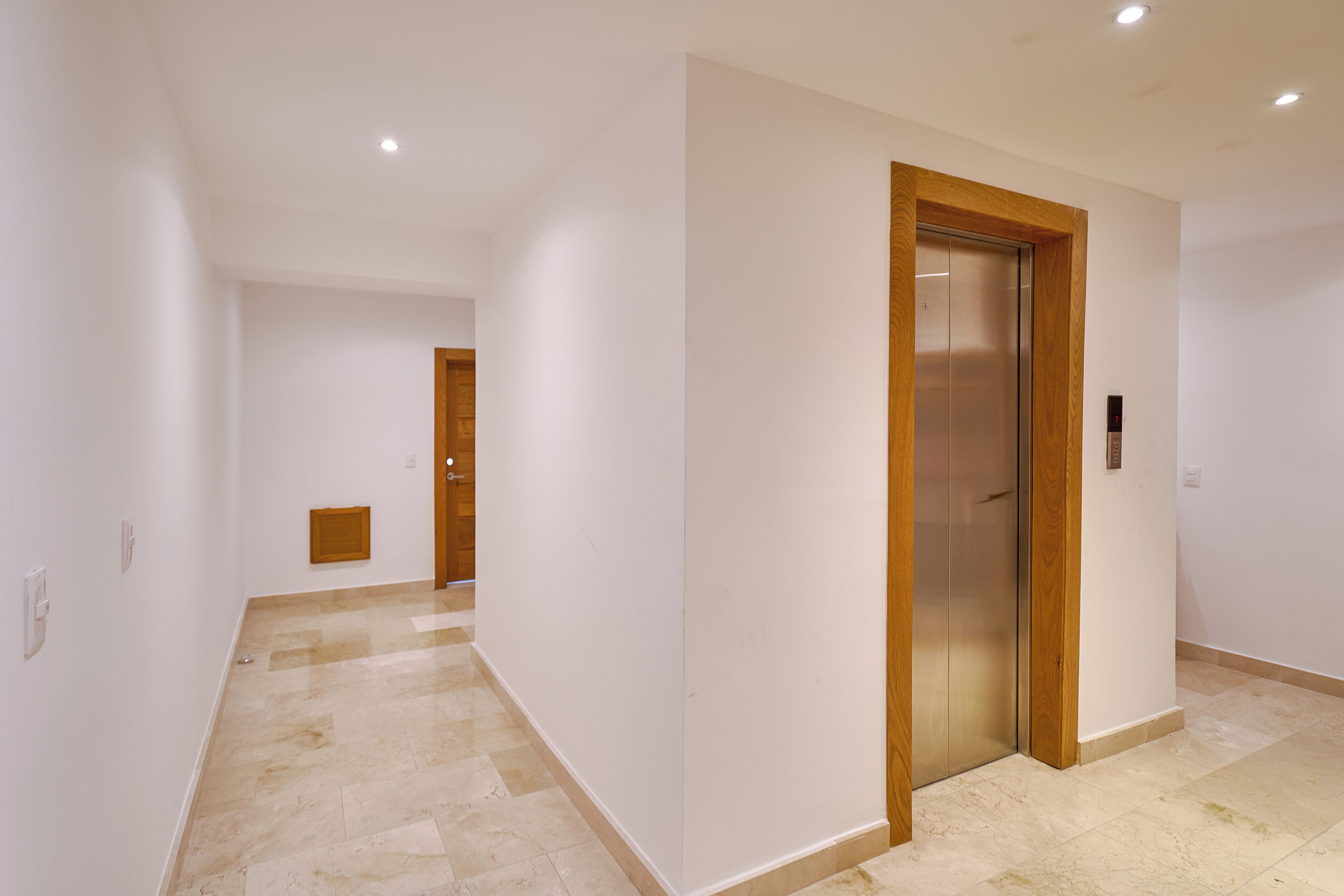 A hallway with a wooden-framed elevator, white walls, and a tiled floor.