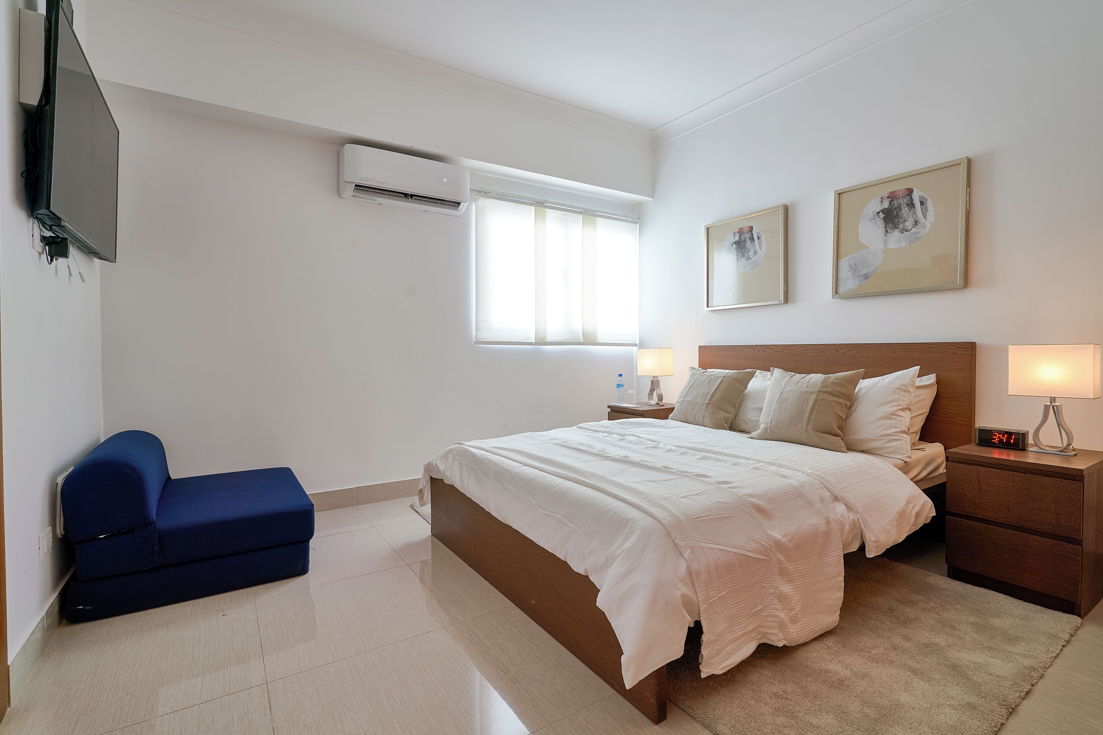 A bedroom with a bed, a blue sofa, a nightstand with a lamp, and two framed pictures on the wall.