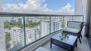 Un balcón con una silla de mimbre y una mesita, con vistas a un paisaje urbano con diversos edificios y áreas verdes.