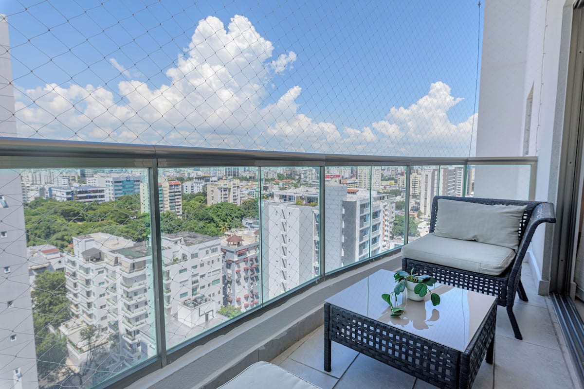 Ein Balkon mit Korbstuhl und kleinem Tisch, mit Blick auf eine Stadtlandschaft mit verschiedenen Gebäuden und Grünflächen.