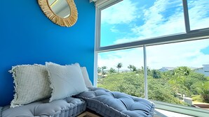 A room with a blue wall, a window with a view of greenery, and a cushioned bench with pillows. - Ed. Nixxus Residence (Ipojuca)