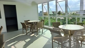 A spacious room with large windows offering a view of a bridge and greenery. There is a round table with chairs and a black door. - Ed. Nixxus Residence (Ipojuca)