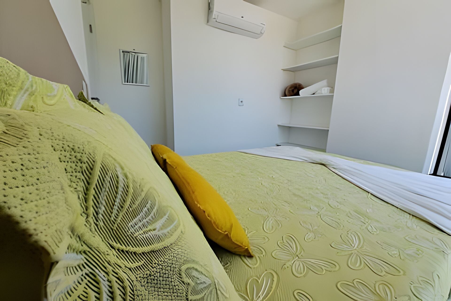 A bedroom with a bed, a yellow pillow, a window, and a white air conditioning unit.