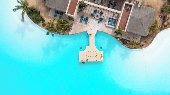 Vue aérienne d’un complexe hôtelier avec une grande piscine turquoise, un bâtiment à toit de chaume et des palmiers.