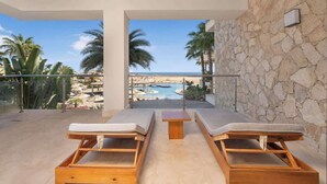 A balcony with two wooden lounge chairs and a small table, overlooking a beach and a pool.