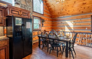 A wooden cabin interior with a dining table, chairs, a black refrigerator, and a stone wall.