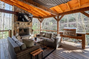 A covered outdoor seating area with a stone fireplace, a television, and a grill. - Slippery Rock-Pet Friendly, Luxury Log Cabin Rental on Creek, Outdoor TV over Fireplace, Hot Tub-Western NC (Topton)