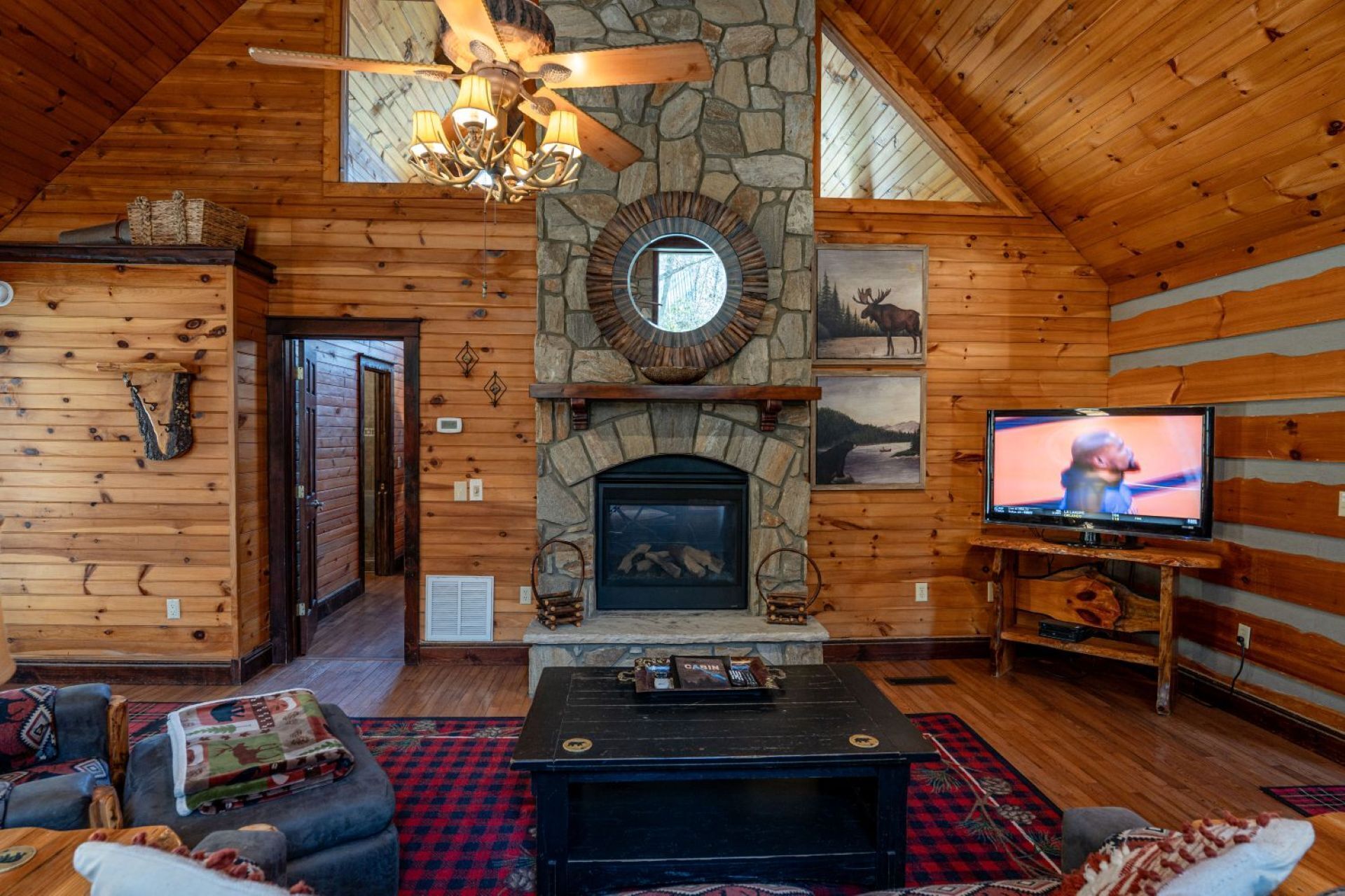 A log cabin interior with a stone fireplace, wooden walls, a ceiling fan, a television, and a cozy seating area.