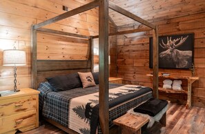 A wooden cabin bedroom with a four-poster bed, a nightstand, a bedside table, a lamp, a framed moose picture, and a shelf with decorative items. - All Decked Out-Pet Friendly, Luxury Log Cabin Rental, High Speed Internet, 4 Decks, Swinging Day Bed, Large Hot Tub-Nantahala NC. (Topton)