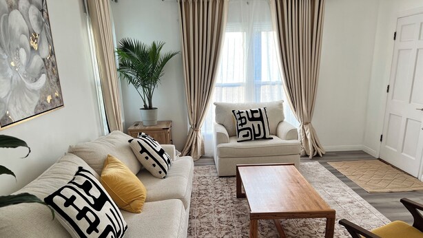 A living room with a beige sofa, a wooden coffee table, and a yellow armchair. There is a potted plant and a painting on the wall.