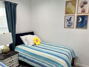 Un letto rifatto con una coperta a righe, un cuscino bianco e un cuscino a forma di fiore giallo. È presente un comodino con una lampada e un cassetto. La stanza ha una finestra con tende blu e opere d'arte appese al muro.