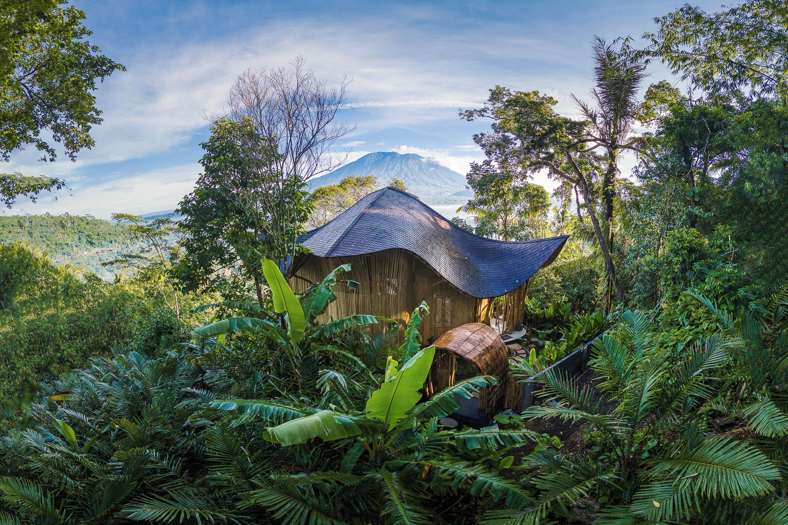 Une construction rustique en bois, dotée d’un toit de chaume, entourée d’une végétation tropicale luxuriante et avec une montagne en arrière-plan.