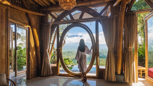 A room with a large circular window, bamboo walls, and a view of a mountainous landscape. - Punduk Villa (Selat)