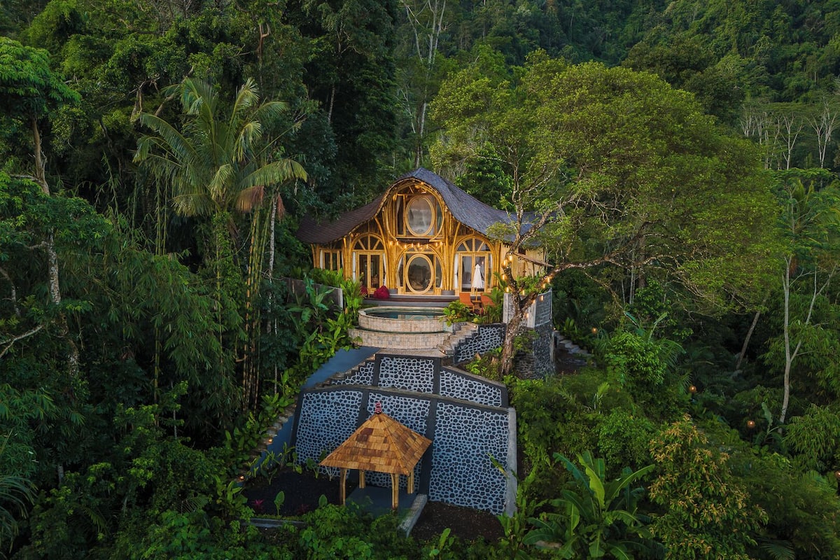 A luxurious villa surrounded by lush greenery, featuring a central courtyard with a stone mosaic floor and a small thatched-roof structure.