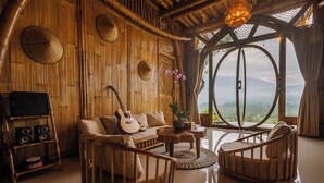 A room with a bamboo wall, a guitar, a sofa, a coffee table, and a view of the mountains. - Punduk Villa (Selat)