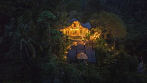 A traditional wooden house surrounded by dense tropical foliage, with a stone base and a thatched roof. - Punduk Villa (Selat)