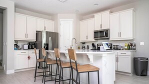 A modern kitchen with white cabinets, a stainless steel refrigerator, a microwave, and a bar area with stools.