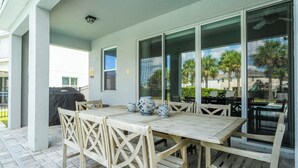 An outdoor patio with a wooden table and chairs, a grill, and a glass door leading to a view of palm trees.