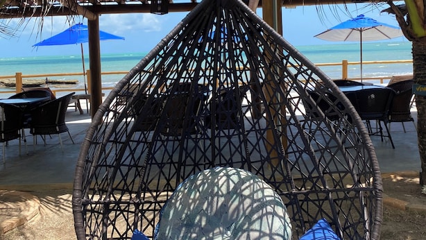 A hanging hammock chair with blue cushions, a wooden ceiling, and a view of the beach and ocean. - Pousada Bangalôs Do Pontal (Japaratinga)