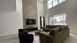A modern living room with a large sofa, a black armchair, and a coffee table. There is a flat-screen TV mounted on the wall and a large window providing natural light.