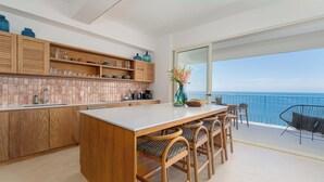 Eine moderne Küche mit Holzausstattung und einer großen Kochinsel, ein Essbereich mit Holzstühlen und ein Balkon mit Meerblick.