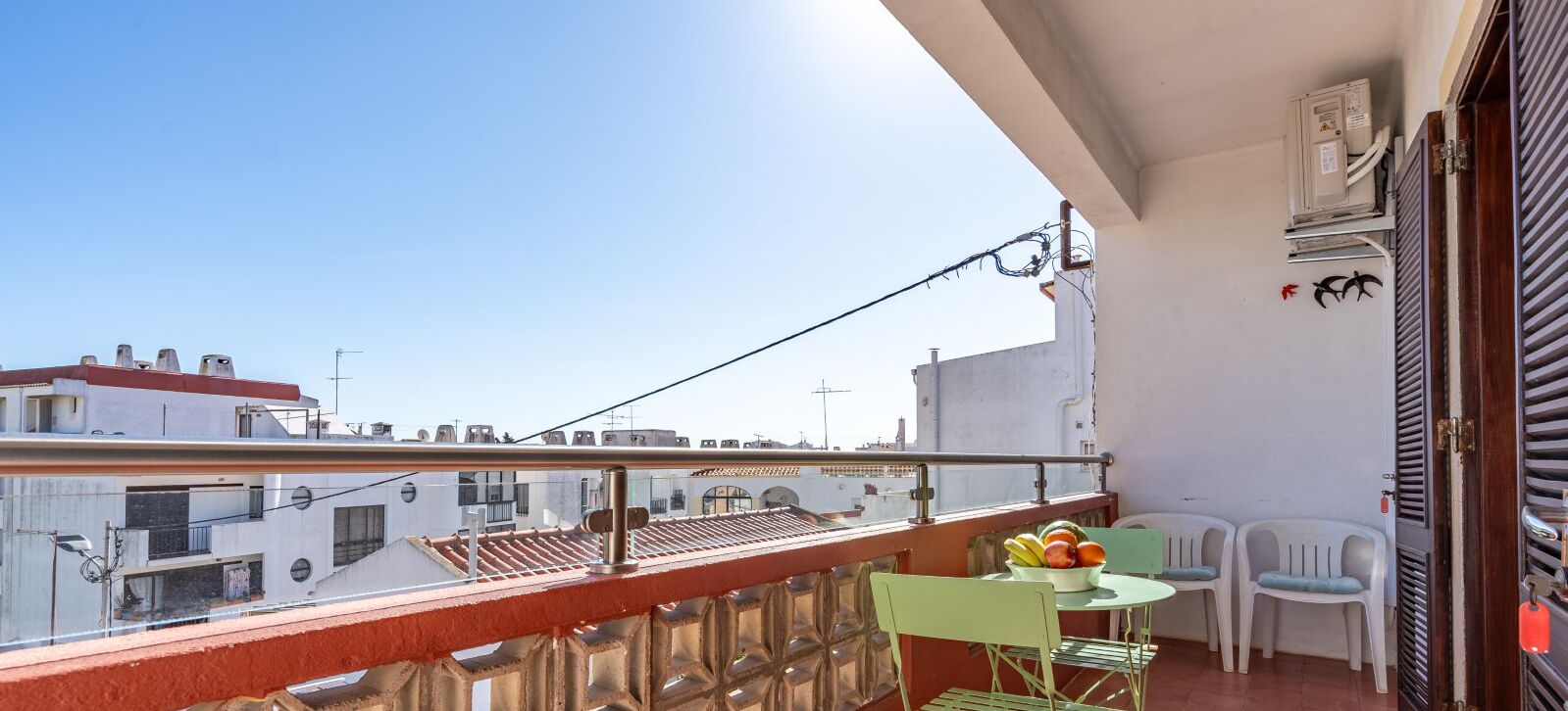 Een balkon met een tafel, stoelen, een schaal fruit en uitzicht op gebouwen.