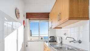 Cocina compacta con baldosas blancas, armarios de madera, una ventana y vista al mar.