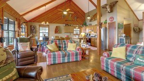 Een ruime woonkamer met een hoge plafond en houten balken, ingericht met lederen fauteuils en geruite banken, en een zichtbare keuken met houten kasten en een koelkast.