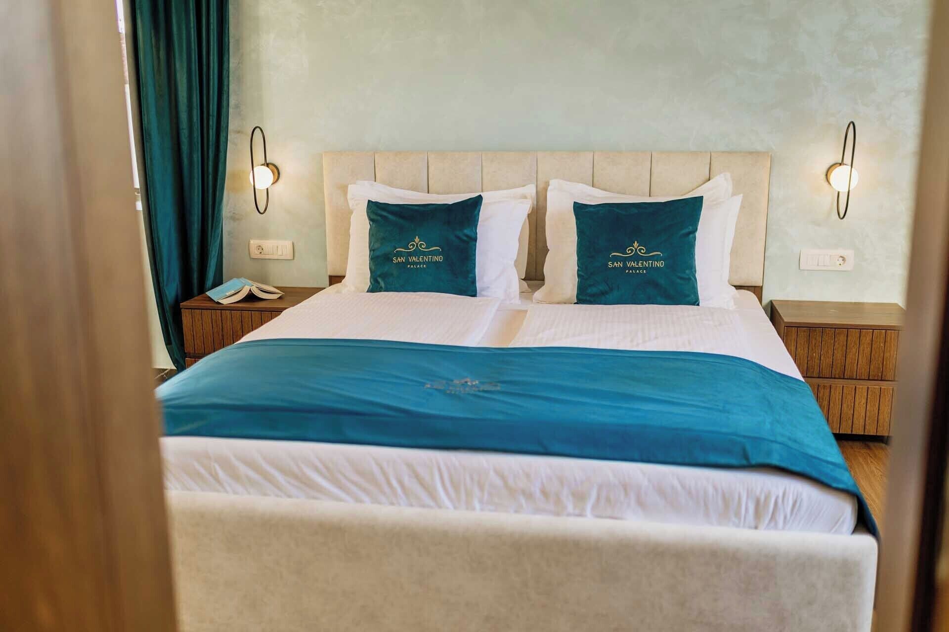 Una cama doble con una colcha blanca y aguamarina, dos almohadas aguamarina con el texto 'San Valentino Palace' y dos mesitas de noche con lámparas.
