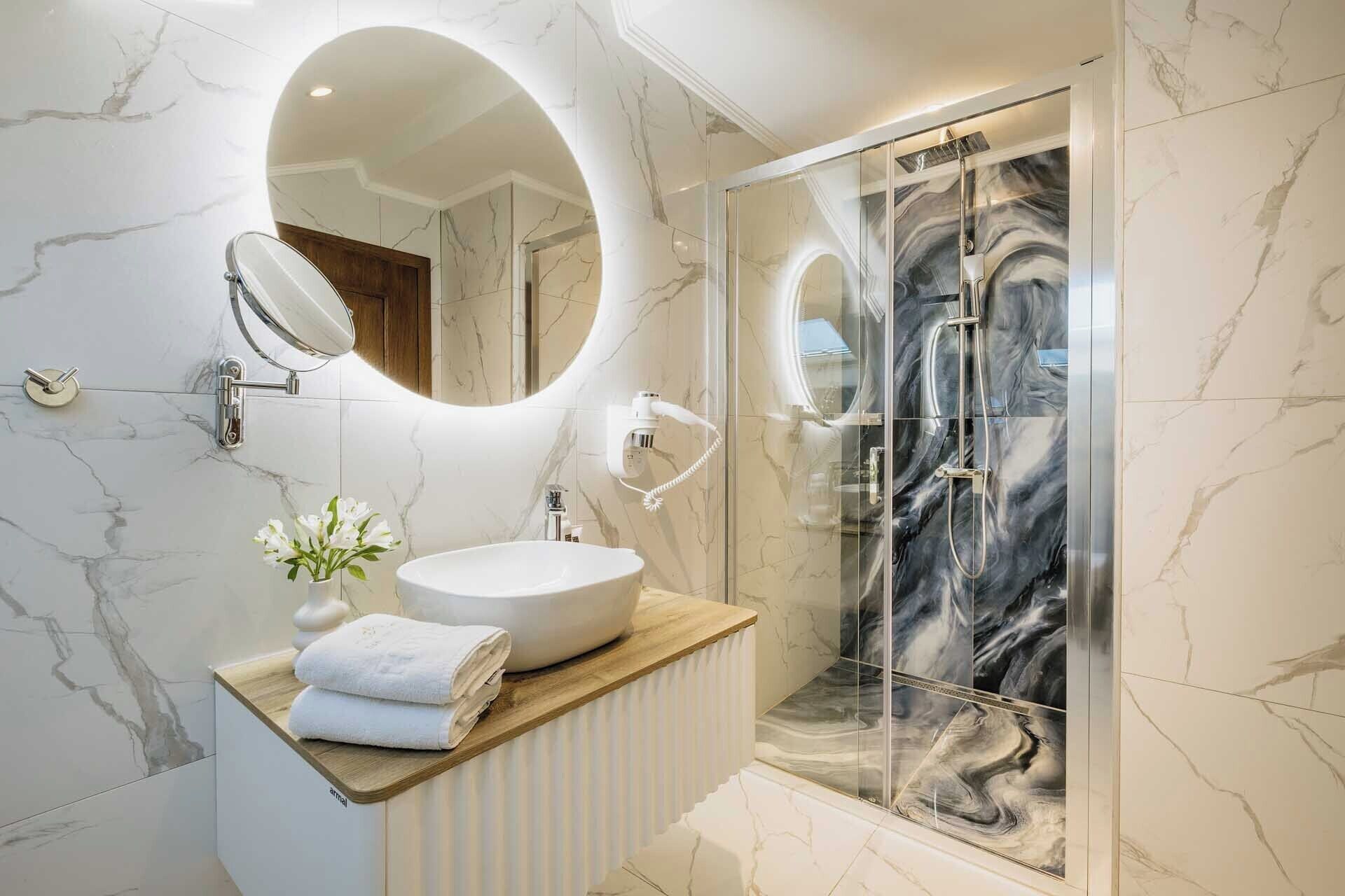 A modern bathroom with a marble vanity, a glass shower enclosure, and a round mirror.