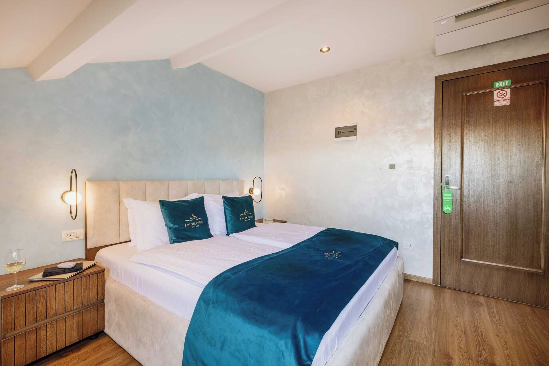 A hotel room with a bed, a wooden nightstand, a glass of wine, and a door with an exit sign.