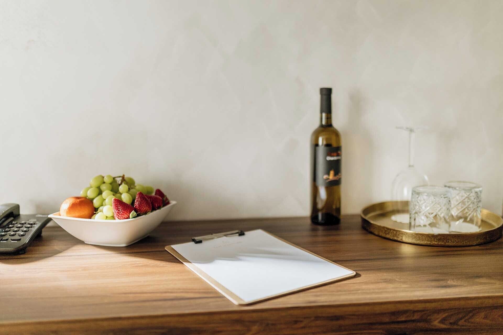 A wooden table with a bowl of fruit, a wine bottle, a clipboard, a calculator, and a glass.