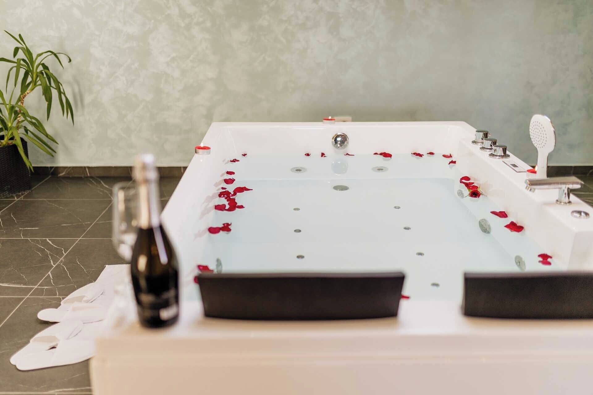 A bathtub with rose petals, a bottle, and slippers on a tiled floor.