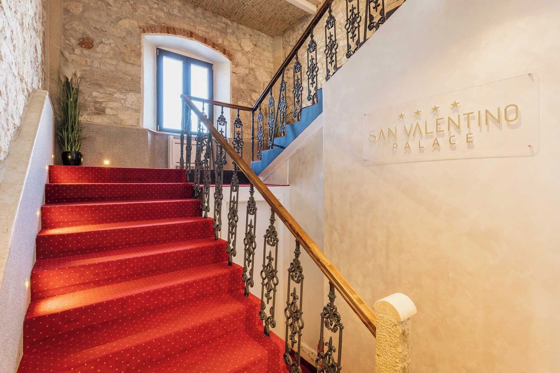 Un escalier recouvert d’une moquette rouge et orné de rampes en métal ouvragé mène à une pièce dont l’un des murs est en pierre et sur laquelle on peut lire l’inscription « Palais San Valentino ».