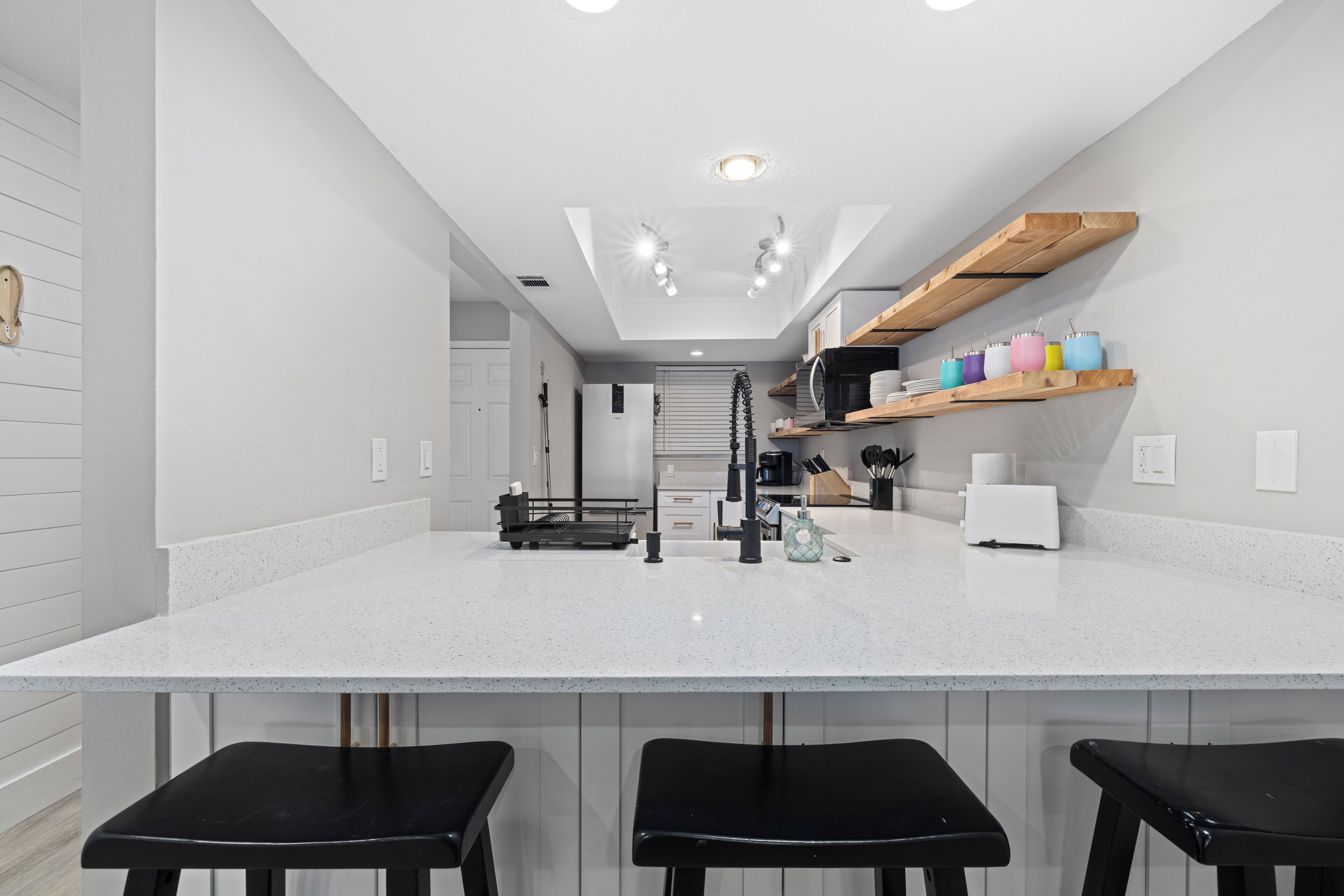 Una cocina moderna con encimera blanca, taburetes negros y estantes de madera.