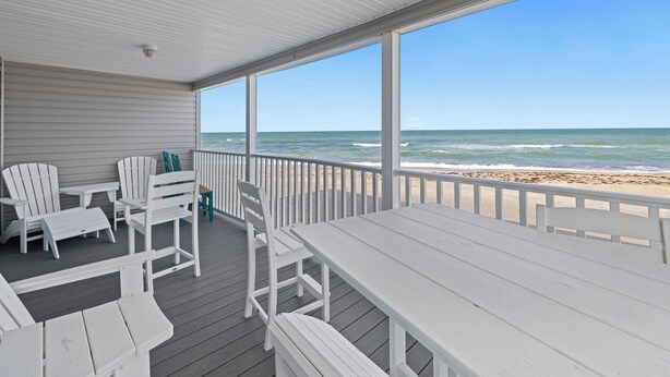 A covered outdoor area with white furniture, including chairs and a table, overlooking a beach.