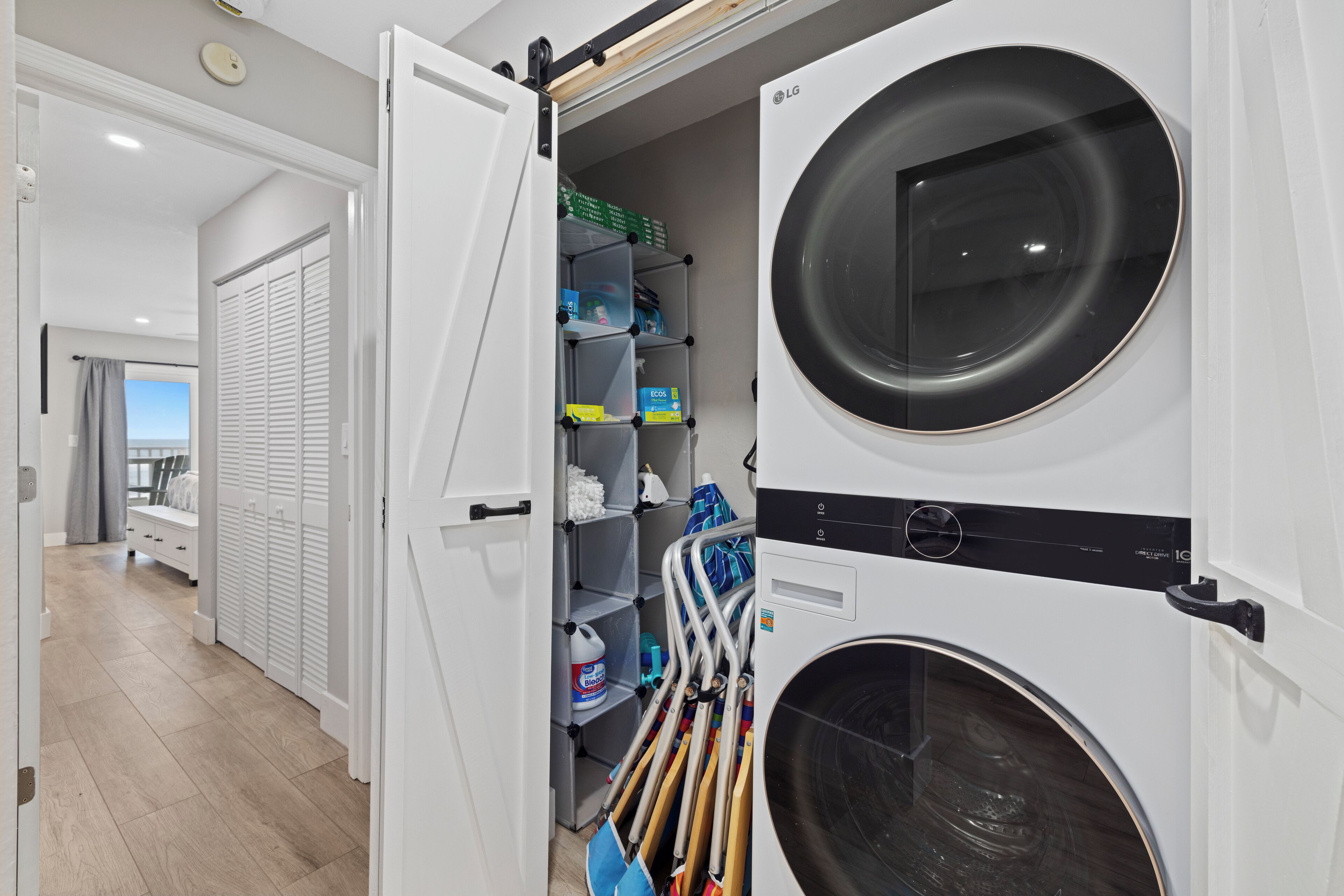 Une buanderie équipée d’un lave-linge à chargement frontal et d’un sèche-linge, de plusieurs étagères où sont rangés les produits de nettoyage, et donnant sur un balcon par une porte ouverte.