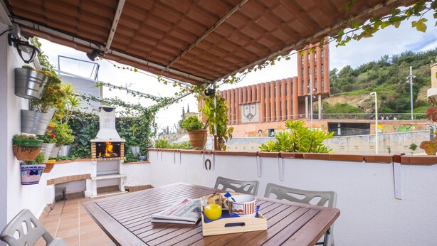 Eine Terrasse im Freien mit einem Holztisch, Stühlen und einem Grill. Übertöpfe mit Pflanzen und ein Blick auf ein Gebäude im Hintergrund.