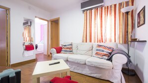 Ein Wohnzimmer mit einem weißen Sofa, bunten Kissen, einem roten Sessel, einem kleinen Tisch und einem Fernseher.