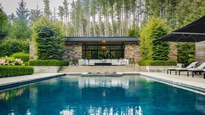 A modern outdoor pool area with a stone and glass structure, surrounded by lush greenery and tall trees. - Stunning 6 bed 5 bath estate on 65 acres with Pool, Hot Tub and Tennis Court! (Caledon Village)