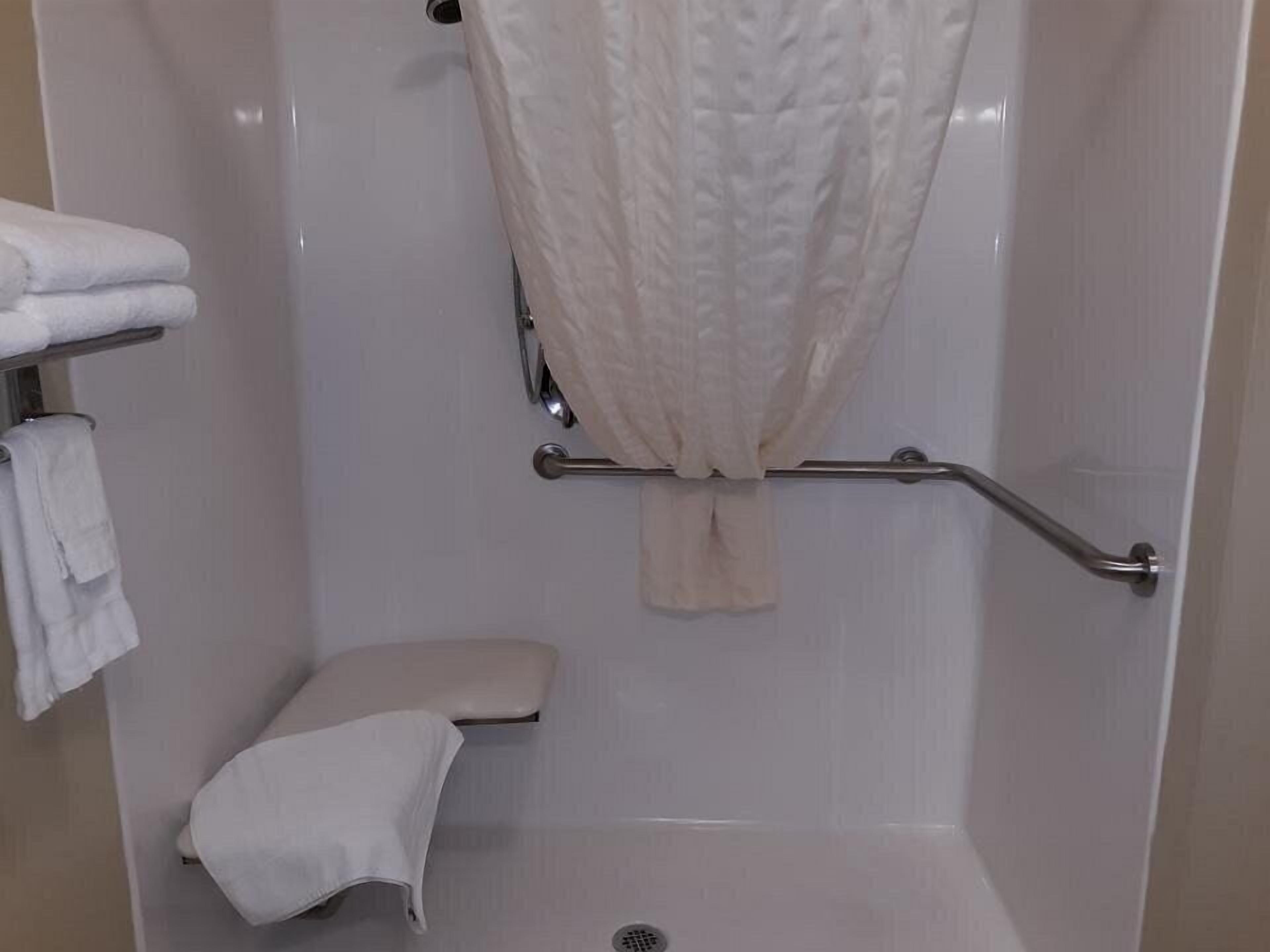 A small, enclosed shower area with a white shower curtain, a grab bar, a bench, and a towel rack with folded towels.