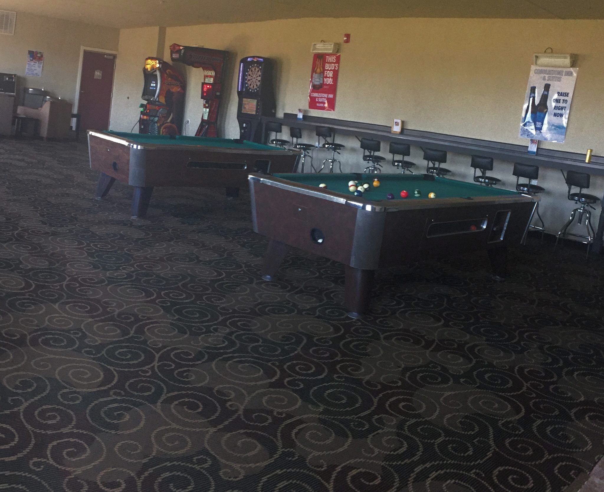 Deux tables de billard dans une pièce dont le sol est recouvert d’une moquette à motifs, ainsi qu’un jeu de fléchettes et une affiche accrochée au mur.