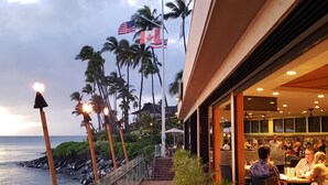 A beachfront restaurant with outdoor seating overlooking the ocean, flags, and palm trees. - Gorgeous Napili Shores studio A/C (Lahaina)
