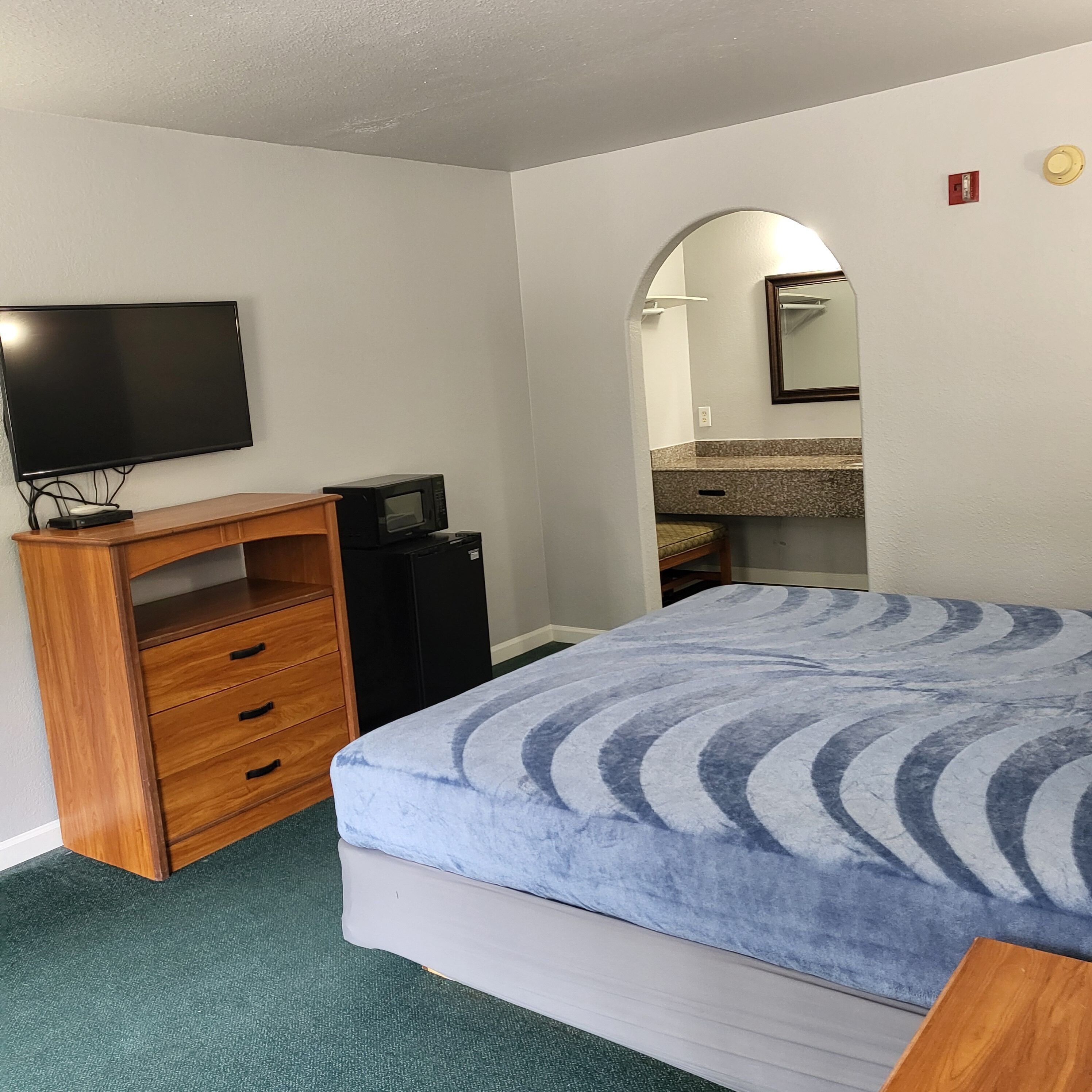 Habitación de hotel con una cama, cómoda de madera, televisor, microondas, refrigerador pequeño, espejo de baño y lavamanos.