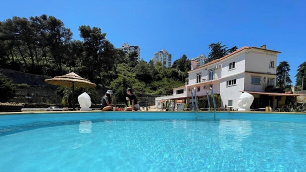 A swimming pool with clear blue water, surrounded by a white building with a red roof, and trees in the background. - Quinta da Villa - House of Wines (Cascais)