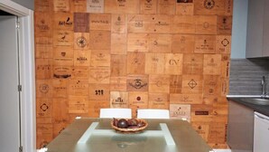 A dining area with a glass table and chairs, a wooden wall with various logos, and a basket of fruits on the table. - Quinta da Villa - House of Wines (Cascais)